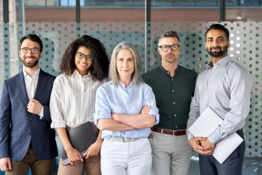 Ein fünfköpfiges Team im Business Outfit steht lächelnd in einem Büro- oder Meetingraum. Es sind zwei Frauen und drei Männer unterschiedlichen Alters und ethnischer Herkunft.