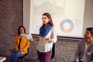 Eine junge Frau steht vor einer Leinwand, auf die Grafiken und Zahlen projeziert sind, und hält einen Vortrag. Ihre Körpersprache ist offen und kommunikativ, um sie herum sitzen weitere Personen, die ihr aufmerksam zuhören.