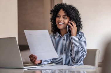 Eine junge Frau mit dunklen Locken sitzt am Schreibtisch, vor ihr ein aufgeschlagener Laptop, und führt ein Telefoninterview für eine Bewerbung. Sie hält einen Zettel mit Notizen in der Hand und lächelt.