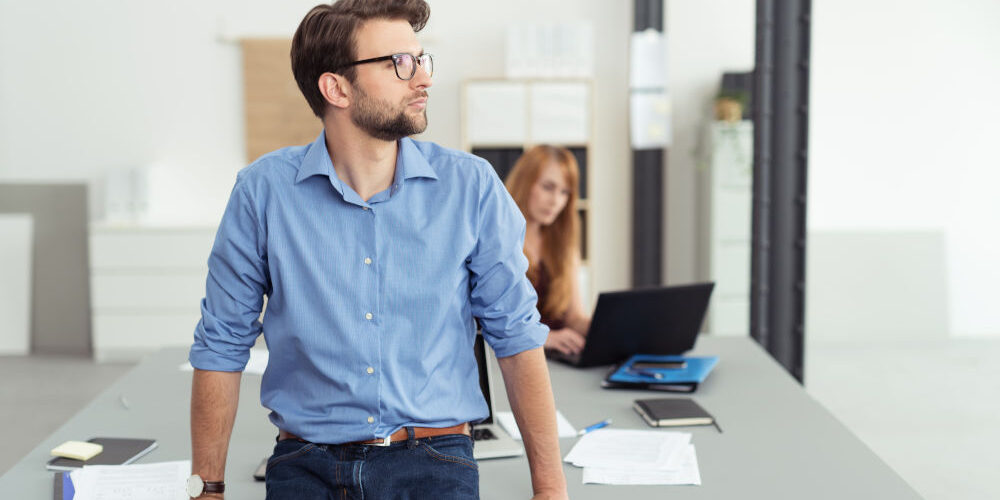 Ein Mann in seinen Dreißigern, gekleidet im Businesslook, steht in einem Büro und denkt über eine berufliche Neuorientierung nach. Er blickt nachdenklich zur Seite, im Hintergrund sitzt eine rothaarige Frau an einem Tisch und arbeitet am Laptop.