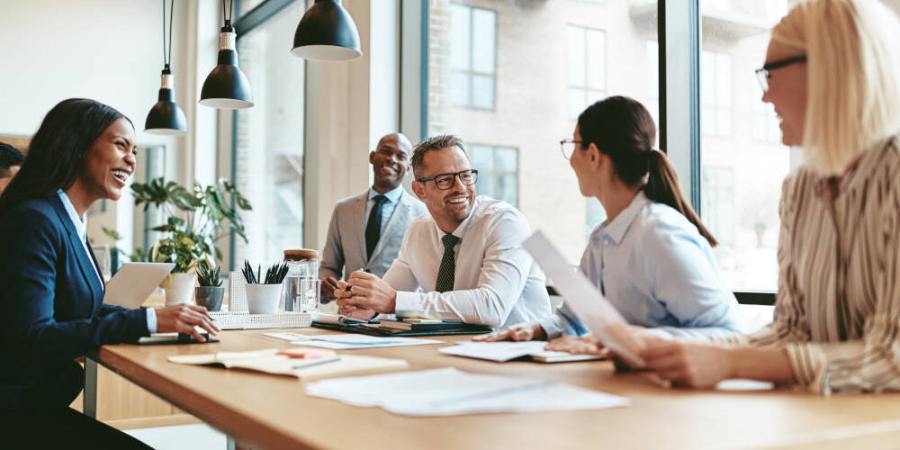 Lernen Sie verschiedene Meeting Arten kennen. Eine Gruppe von Menschen sitzt in einem hellen Raum mit großen Fenstern um einen großen Besprechungstisch herum.