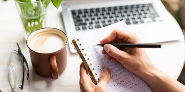 Female hands holding notebook and pen while making working notes by table in cafe and having fresh cappuccino