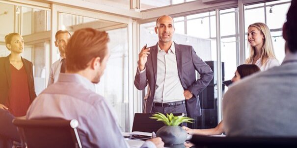 Ein mittelalter Mann im Anzug steht in einem lichtdurchfluteten Raum an einem Konferenztisch, um ihn herum sitzen und stehen weitere Personen und hören ihm zu. Das Bild illustriert, welche Fähigkeiten es braucht, um Teamleiter zu werden: Erfahrung, soziale und fachliche Kompetenz.