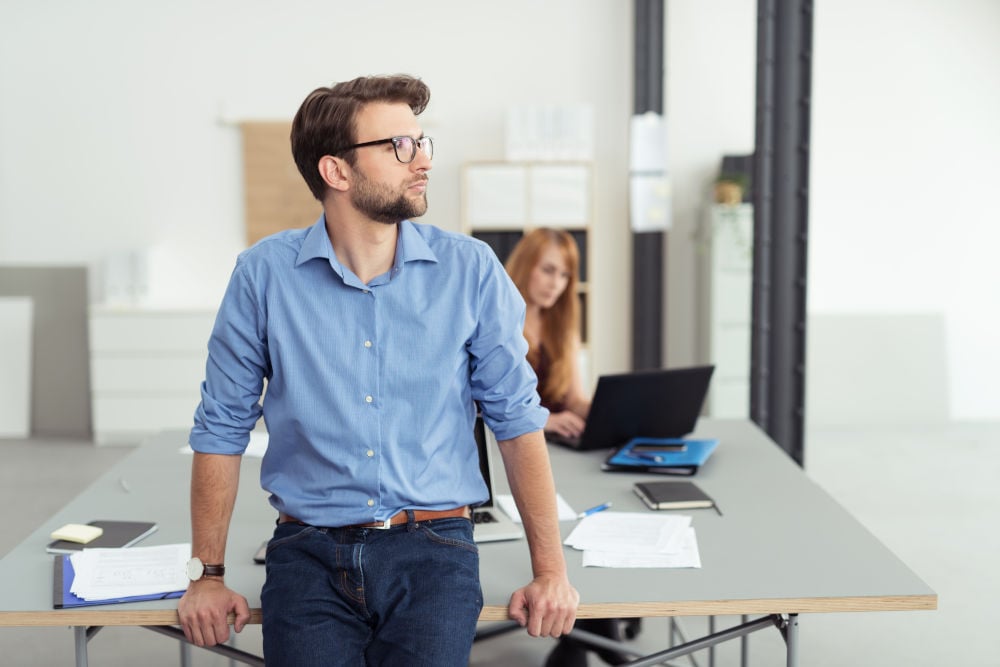 Ein Mann in seinen Dreißigern, gekleidet im Businesslook, steht in einem Büro und denkt über eine berufliche Neuorientierung nach. Er blickt nachdenklich zur Seite, im Hintergrund sitzt eine rothaarige Frau an einem Tisch und arbeitet am Laptop.
