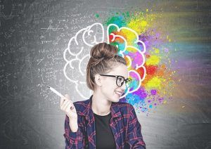 Eine lächelnde junge Frau mit hellen Haaren und Brille sitzt mit einem Stift in der Hand vor einem grauen Hintergrund. Hinter ihrem Kopf ist mit weißen Linien ein Gehirn an die Wand gezeichnet, die rechte Gehirnhälfte ist mit bunten Farben ausgemalt, um das Thema Kreativitätstechniken zu illustrieren. Sie lacht und blickt zur rechten Seite.