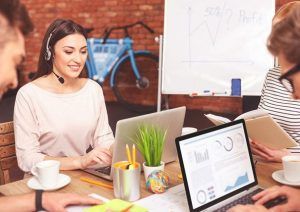 Eine junge dunkelhaarige Frau sitzt zusammen mit mehreren Personen, die jedoch nur teilweise zu sehen sind, in einem Büro und nimmt an einer Telefonkonferenz teil. Sie trägt Kopfhörer und schaut lächelnd auf einen Laptop.
