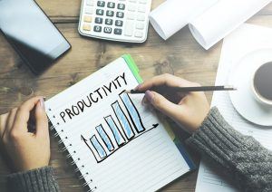 Eine Person, von der man nur die beiden Arme und Hände sieht, zeichnet ein Säulendiagramm mit stetig größer werdenden Säulen auf einen Block, darüber steht das englische Wort Productivity. Der Block liegt auf einem dunklen Holztisch, daneben liegen ein Taschenrechner, Papiere und eine volle Kaffeetasse. Das Bild soll das Thema Produktivität steigern illustrieren.