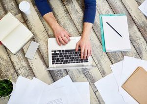 Ein Mann, von dem wir nur die Arme und Hände sehen, sitzt an einem Holtisch und tippf auf einem Laptop. Er fertigt ein Protokoll nach einer der im Artikel genannten Protokollarten an. Um den Laptop herum liegen verschiedene Papiere, Bücher, Stifte, außerdem stehen dort ein Coffe-to-go-Becher und eine kleine Pflanze.