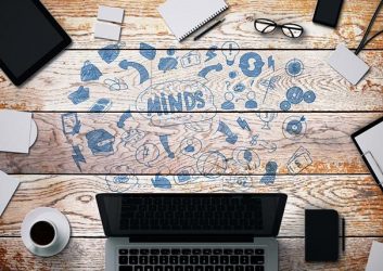 Wir gucken von oben auf einen Holztisch, der in der Mitte voll gekritzelt ist mit Sketchnotes: Ein Gehirn, Pfeile und andere visuelle Elemente. Drum herum stehen ein Laptop, eine Kaffeetasse, ein Notizblock, ein Handy und eine Brille.