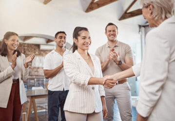 So geht Wertschätzung für Mitarbeiter: Das Foto zeigt eine Gruppe lächelnder Menschen in einem hellen Büro, die in einem Halbkreis um eine junge, dunkelhaarige Frau stehen, die einer älteren Frau mit graublonden Haaren lachend die Hand schüttelt.