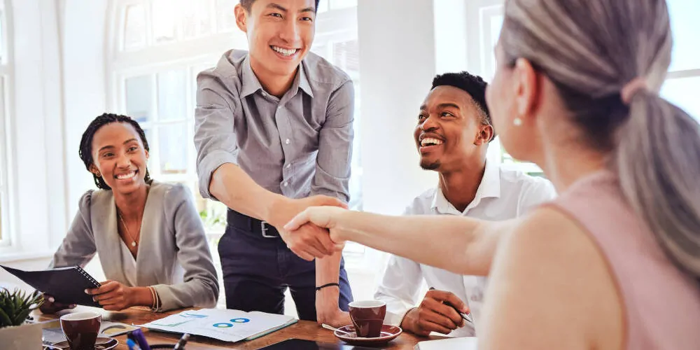 Ein Mitarbeiter erhält ein Lob für gute Arbeit, seine Vorgesetzte reicht ihm die Hand, er lächelt, neben ihm sitzen zwei weitere Kollegen, ein Mann und eine Frau, die ebenfalls lächeln.