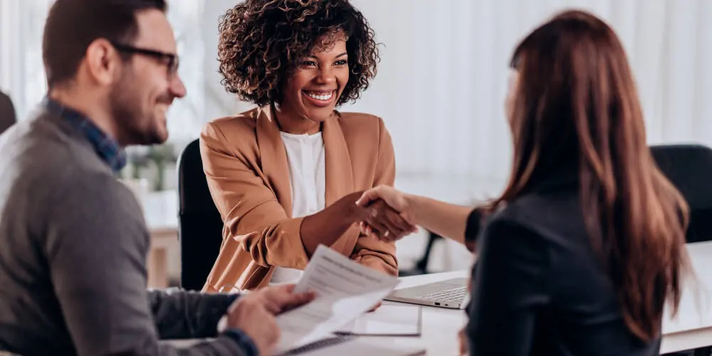 Eine junge Frau mit langen braunen Haaren sitzt beim Vorstellungsgespräch einer ein wenig älteren Schwarzen Frau gegenüber, sie schütteln sich die Hände, die ältere Frau lächelt, die junge Frau ist nur von hinten zu sehen. Daneben sitzt ein junger Mann und halt das Referenzschreiben der jungen Frau in der Hand.