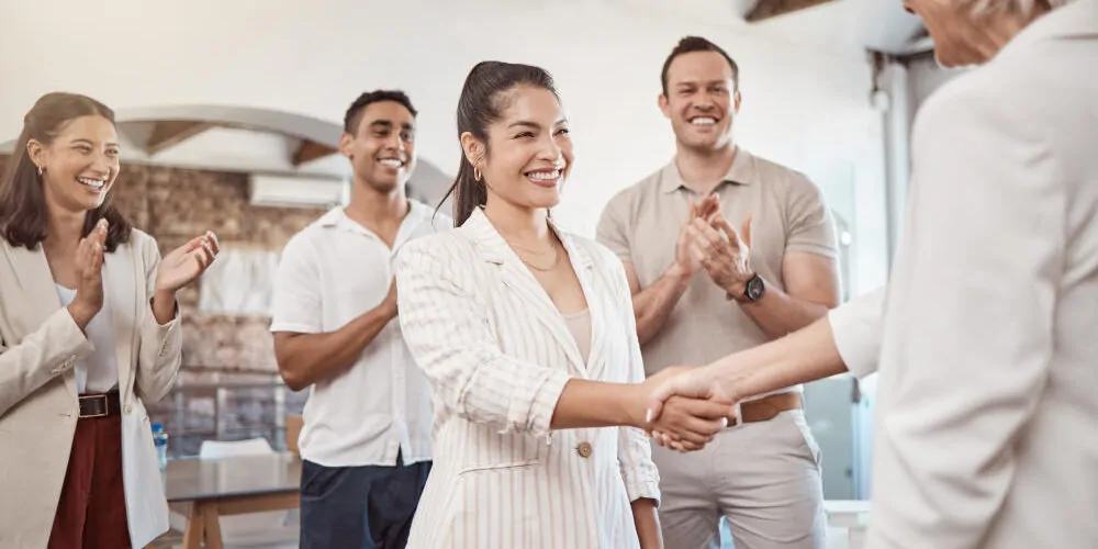 So geht Wertschätzung für Mitarbeiter: Das Foto zeigt eine Gruppe lächelnder Menschen in einem hellen Büro, die in einem Halbkreis um eine junge, dunkelhaarige Frau stehen, die einer älteren Frau mit graublonden Haaren lachend die Hand schüttelt.