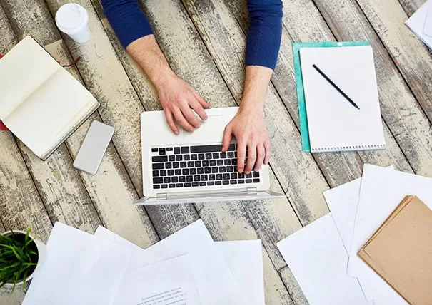 Ein Mann, von dem wir nur die Arme und Hände sehen, sitzt an einem Holtisch und tippf auf einem Laptop. Er fertigt ein Protokoll nach einer der im Artikel genannten Protokollarten an. Um den Laptop herum liegen verschiedene Papiere, Bücher, Stifte, außerdem stehen dort ein Coffe-to-go-Becher und eine kleine Pflanze.